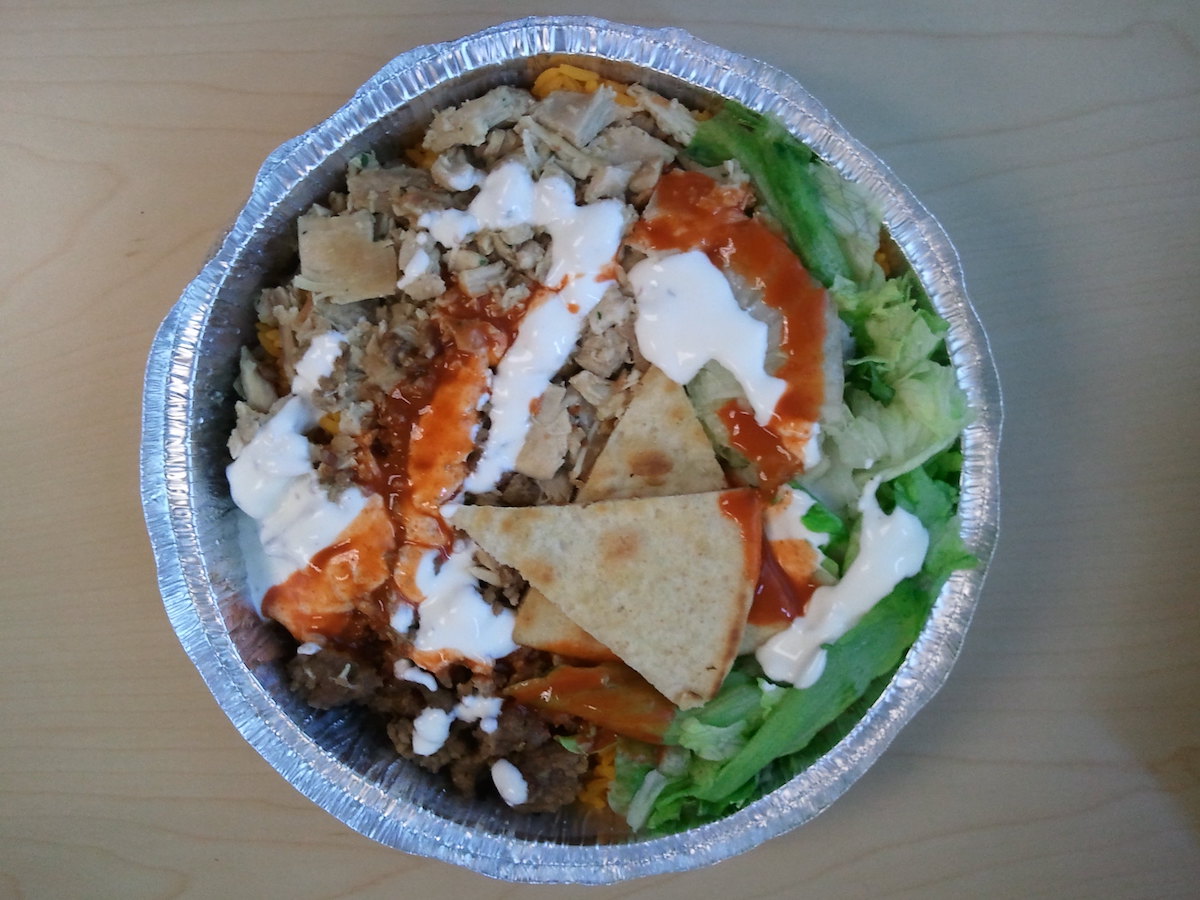 Un recipiente redondo de aluminio lleno de pollo cocido, arroz, lechuga y pita cubierto con lloviznas de salsa roja y blanca hecho por Chicken & Rice Guys, un vendedor ambulante de comida en Boston.