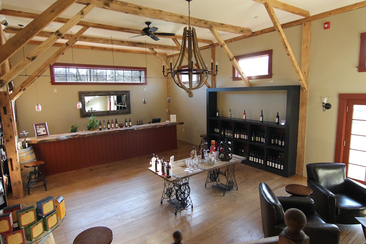 Toma de gran angular de una sala de degustación de bodegas en massachusetts con una barra larga cubierta de botellas de vino, una mesa en el medio de la sala con copas de vino y botellas de vino para degustación