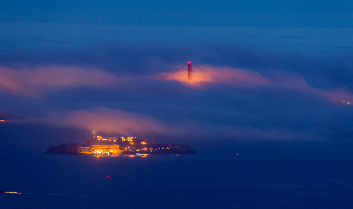 Fotografía de la isla de Alcatraz en San Francisco de noche, con la parte superior del puente Golden Gate sobresaliendo de la niebla
