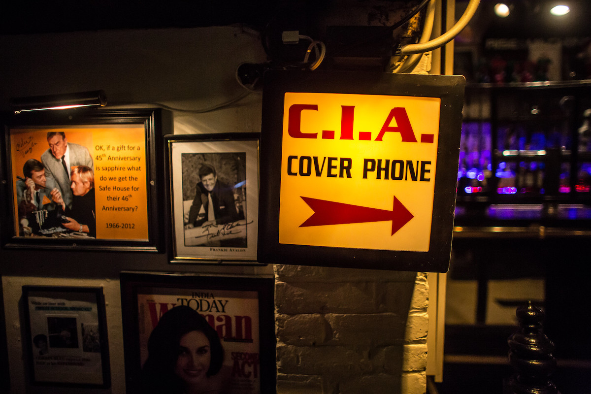 Una pared en un restaurante espía cubierto de fotos y notas, con un gran letrero luminoso que dice "Teléfono de tapa de la CIA" 