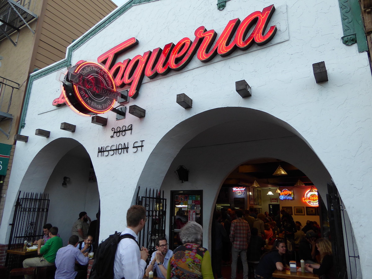 Un restaurante mexicano con una fachada pintada de blanco, dos arcos y un gran letrero de neón rojo que dice "La Taqueria"