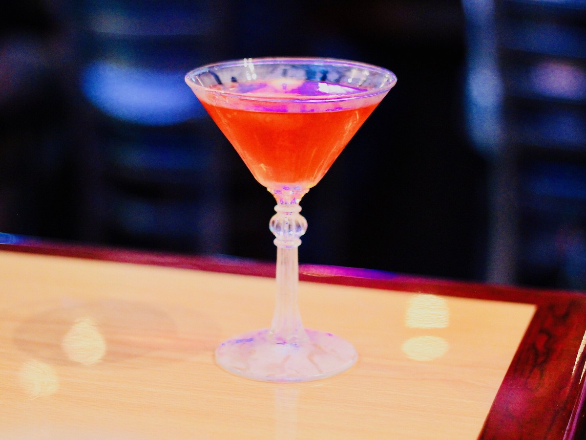 Cerca de un cóctel rojo brillante en una copa de martini sentado encima de una barra de madera