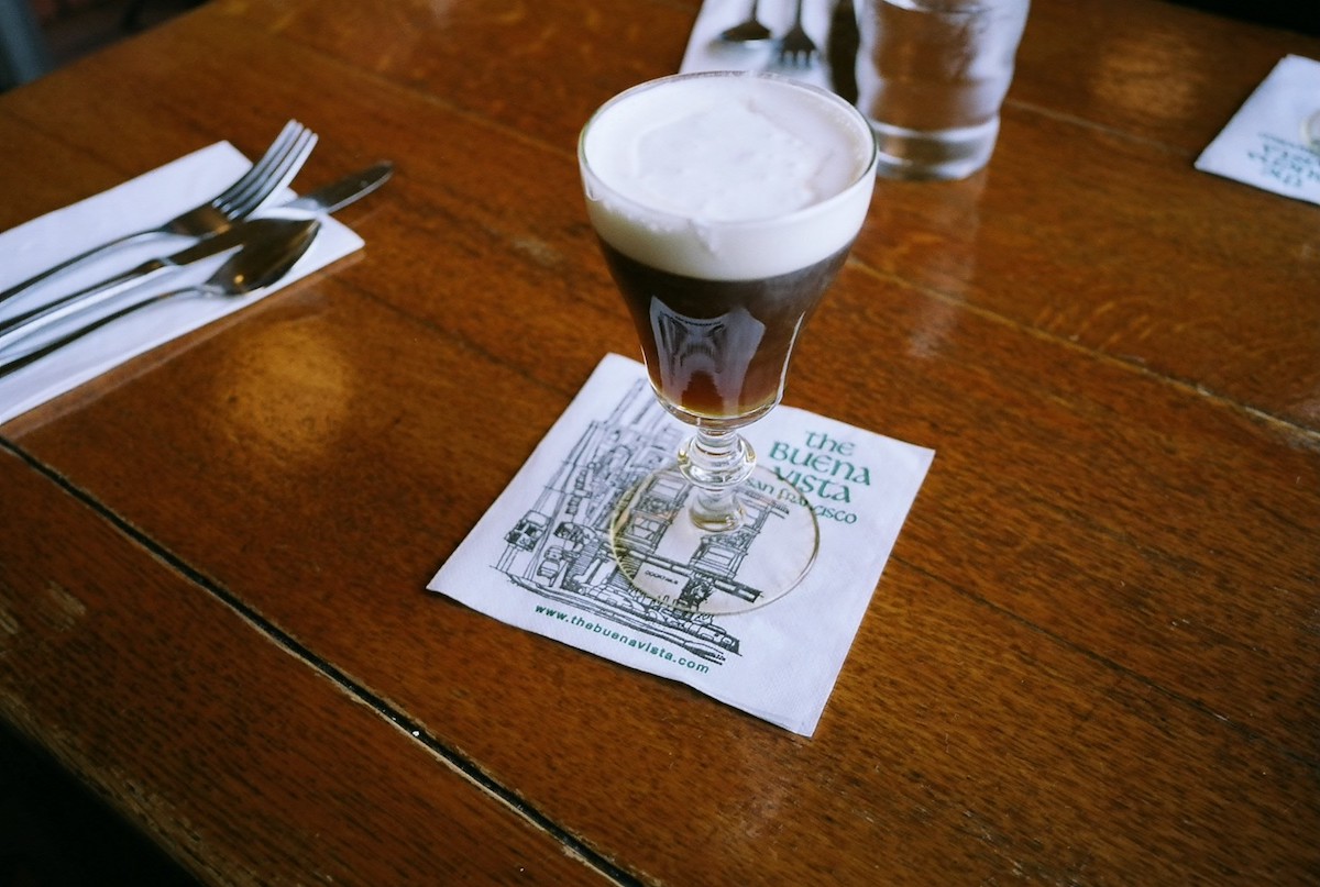 Una mesa de madera pulida con una taza transparente de café irlandés espumoso, encima de una servilleta blanca que dice "The Buena Vista".