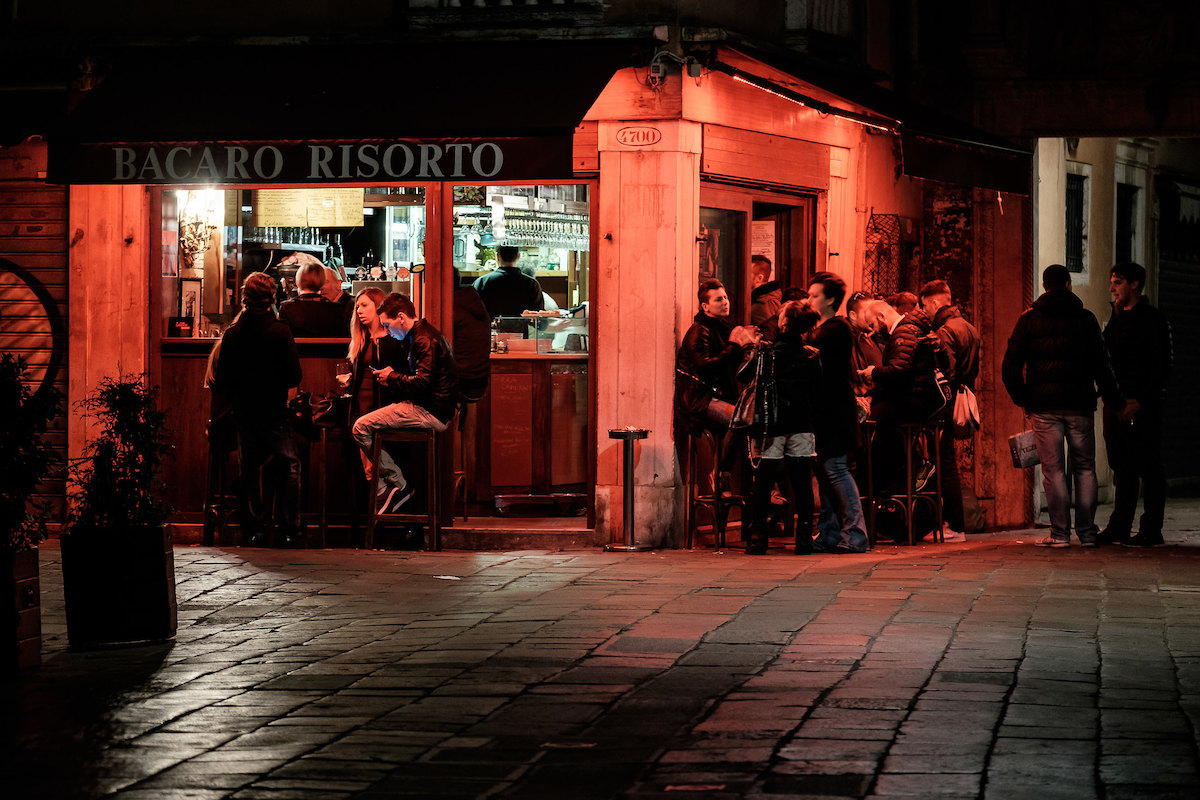 Una foto de un pub en la esquina por la noche en Venecia, iluminado por luces rojas. La gente se sienta y se pone de pie en las mesas, disfrutando del vino y los bocadillos.