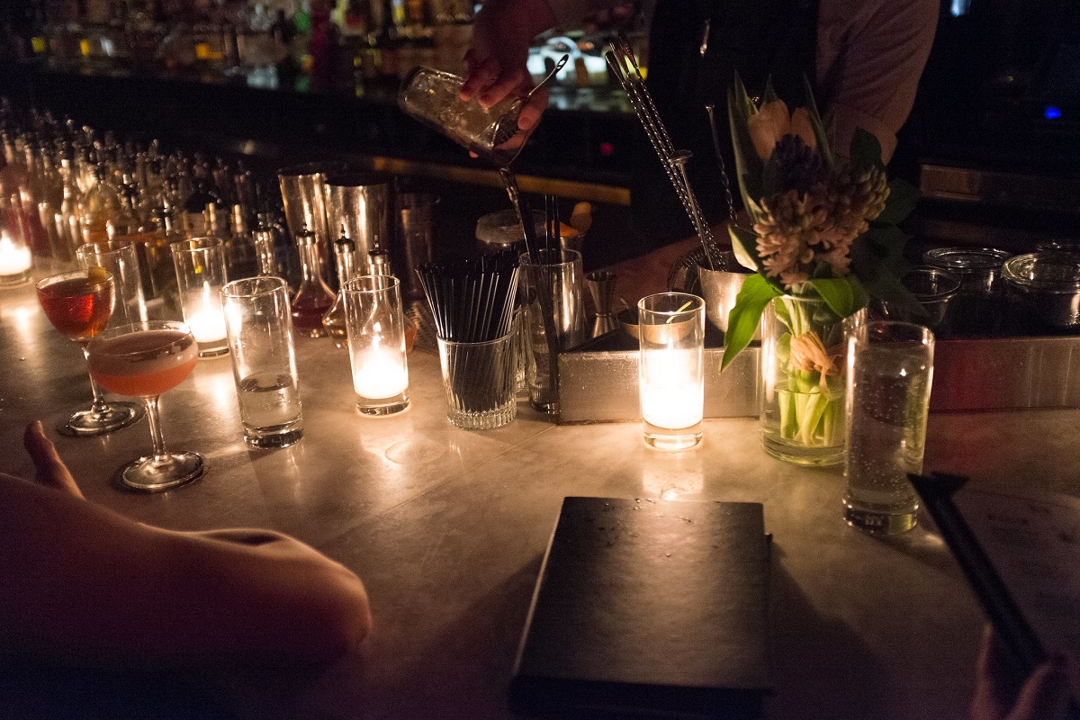Un bar de cócteles sin iluminación con un hombre mezclando cócteles en una coctelera detrás del mostrador.