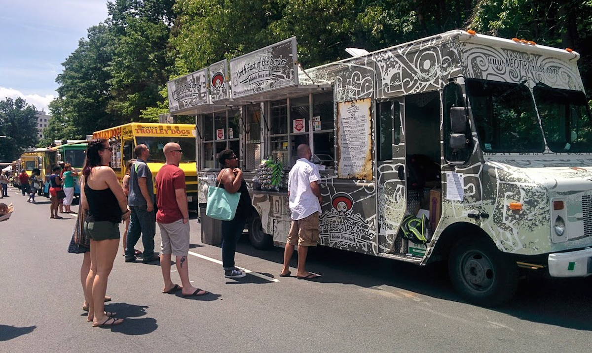 La gente hace fila para ordenar en su camión de comida en el festival de camiones de comida de Boston. El camión está estacionado en la calle y docenas de otros camiones de comida están estacionados en una fila que va al fondo.