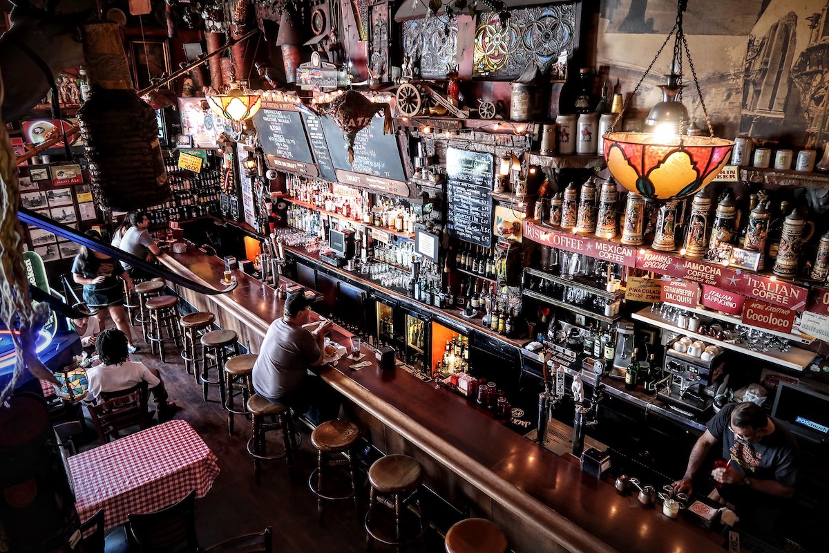 La gente se sienta en el bar de Tommy's Joynt, un famoso restaurante y bar en San Francisco. El bar está repleto de botellas y el interior es todo de madera oscura y pulida.