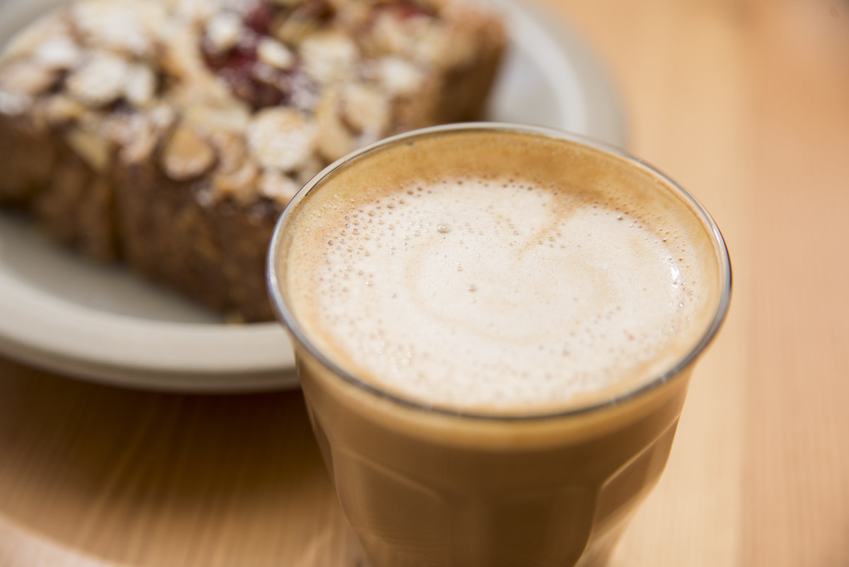 muy cerca de una bebida de café con leche espumada en un vaso transparente, con un pastel en un plato blanco en el fondo
