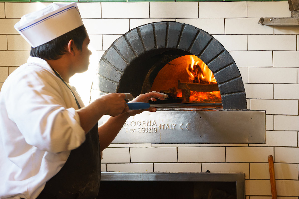 Un chef que lleva una chaqueta blanca y un delantal de cáscara de pizza hornea una pizza en un auténtico horno de pizza de leña de Italia