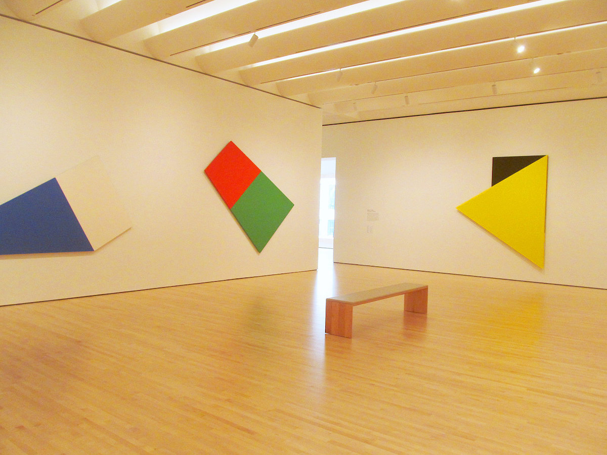 Disparo dentro de una galería en SFMOMA. La habitación tiene paredes blancas y pisos de madera, con un banco de madera en el medio de la habitación y arte abstracto en las paredes hecho de formas brillantes.