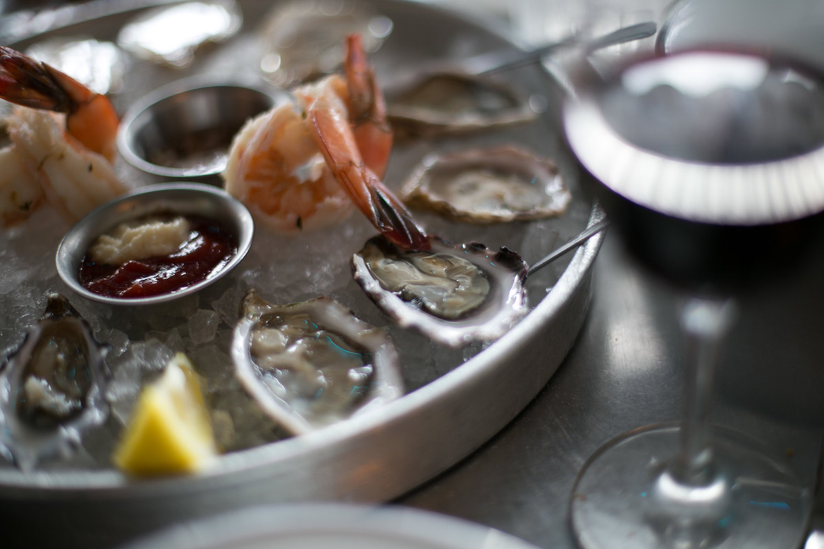 muy cerca de un plato de metal con ostras en hielo, latas de salsa de metal y gambas hervidas. Una copa de vino tinto se ve borrosa en primer plano.