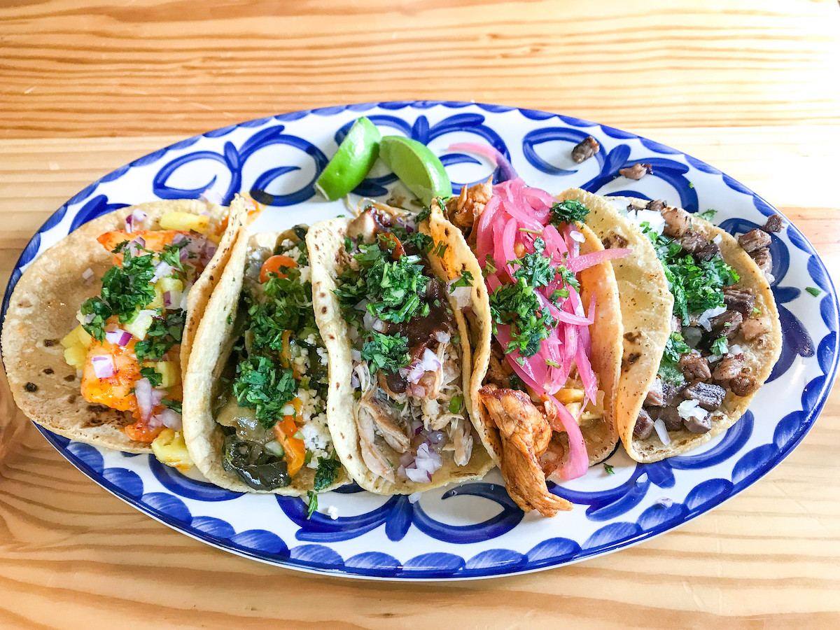 Hay una mesa de madera con un plato pintado de azul y blanco encima con cinco tacos y gajos de lima.