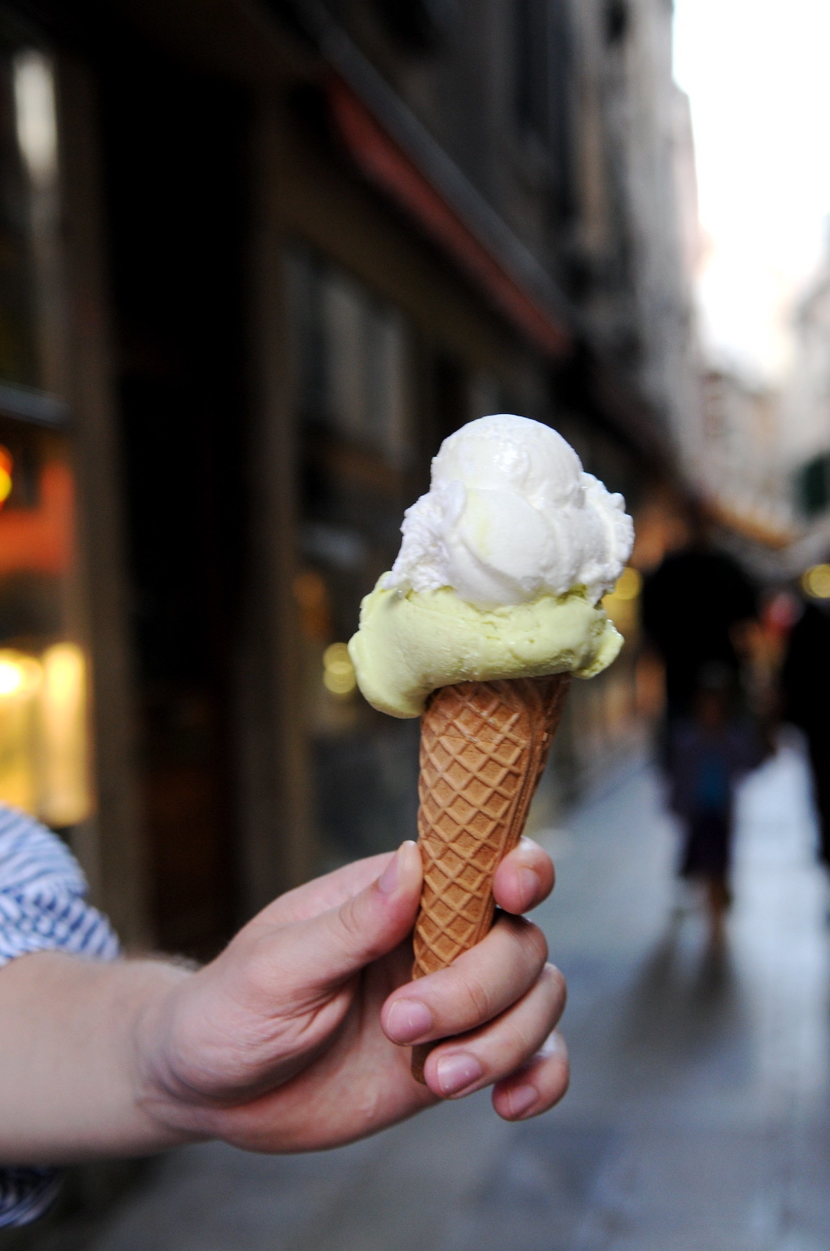 primer plano de la mano de una persona sosteniendo un pequeño cono de gofre con dos bolas de helado, una verde claro y otra blanca, con la calle de Venecia al fondo 