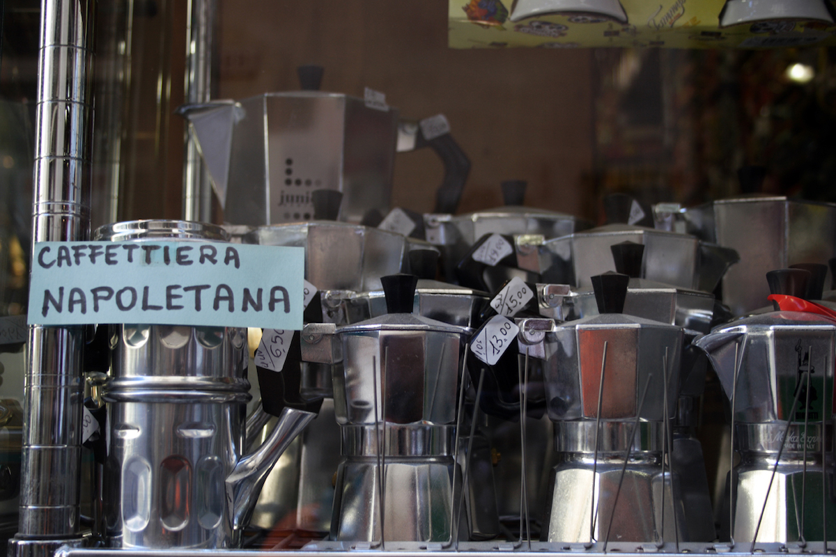 Una exposición de cafeteras en casa en Italia. Hay una olla de cuccuma de estilo napolitano a la derecha con un letrero de papel que dice "caffeteria napoletana", el resto del estante está lleno de ollas Moka brillantes