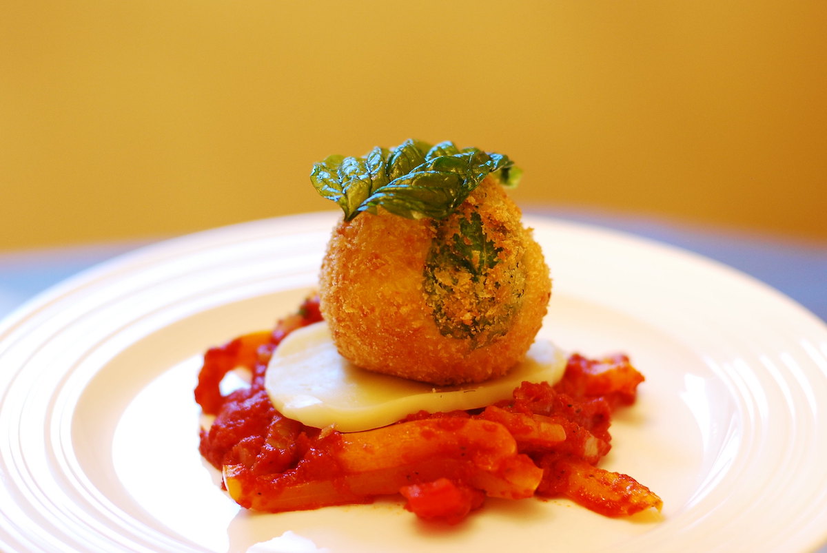 Un arancini cerrado, una pequeña bola de risotto empanizada y frita, cubierta con tomates picados y una rodaja de queso mozzarella.