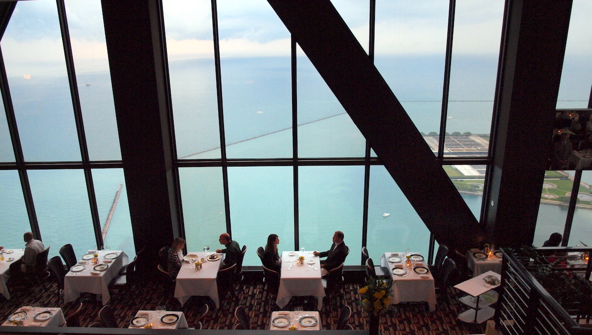 Tomada desde el interior de la Signature Room en Chicago, con ventanas del piso al techo con vista al agua. La gente se sienta en mesas con manteles blancos y disfruta de su comida.