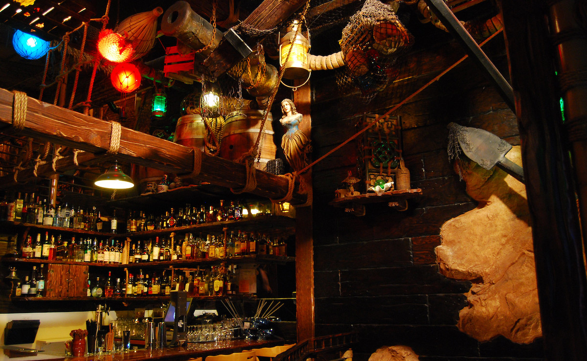 Dentro de la coctelería tiki Smuggler's Cove en San Francisco. El espacio está rodeado por paneles de madera oscura y detrás de la barra hay estantes llenos de botellas de licor.