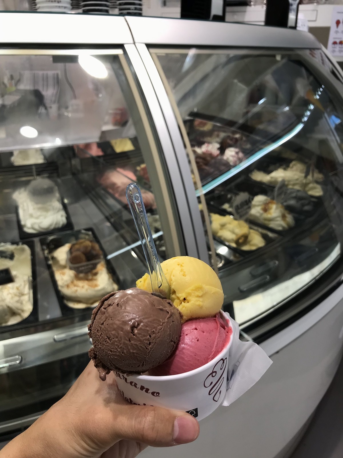 Una persona sostiene una taza blanca con tres bolas de helado, una marrón, una amarilla y una rosa, en una heladería en Venecia.