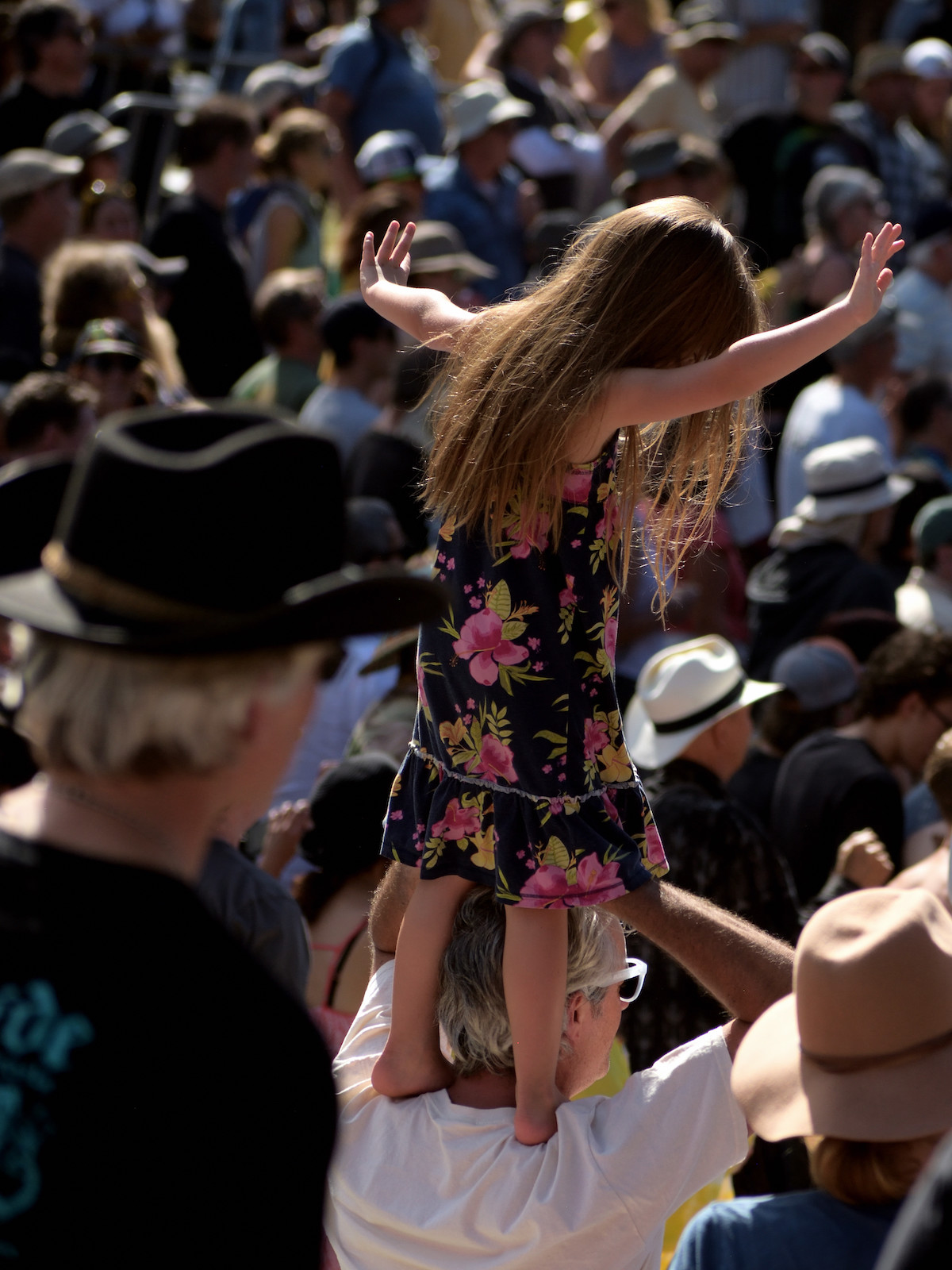 Una niña con un vestido negro con flores rosas en los hombros de un hombre se encuentra entre una gran multitud en un festival de música de verano en San Francisco.