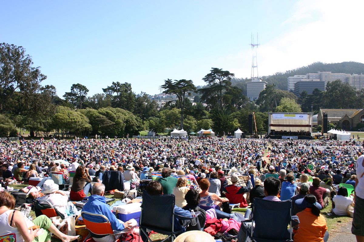 Una gran multitud de personas se sientan en sillas de jardín repartidas por un enorme césped, frente a una representación teatral de Opera in the Park en San Francisco.