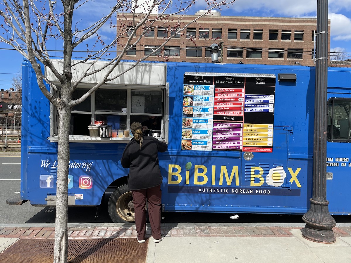 una persona se para en la ventana de pedidos de un camión de comida azul en Boston. Letras amarillas brillantes dicen "Bibim Box" en el costado del camión.