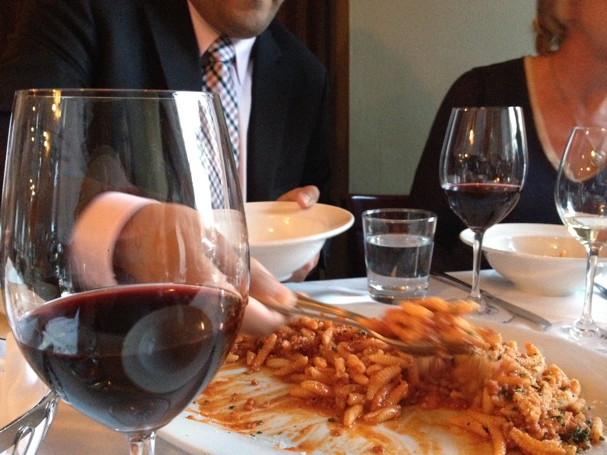 Dos personas se sientan en una mesa con vino tinto en copas de vino, compartiendo un plato de pasta