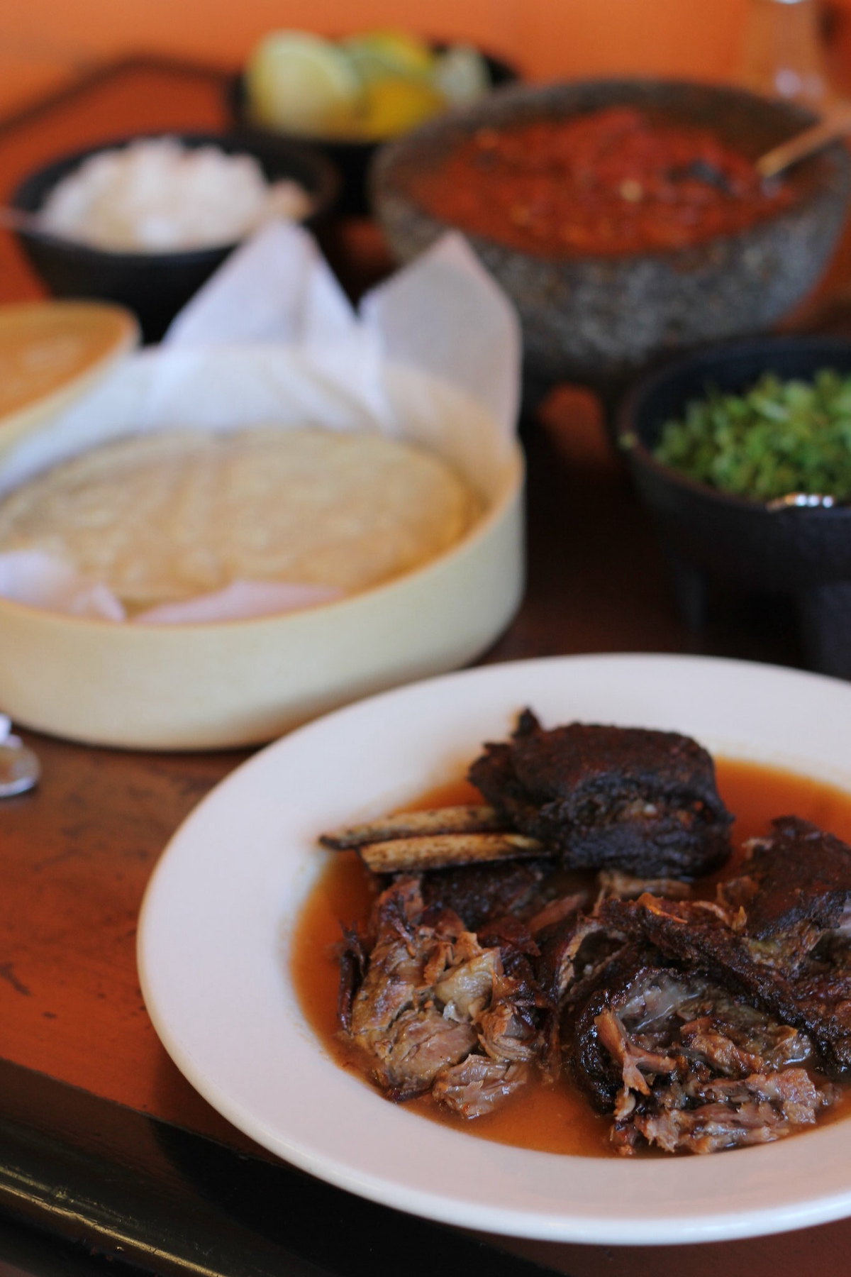 Cierra la mesa en Birrieria Zaragoza en Chicago. Hay carne de cabra guisada en un plato blanco en primer plano, con salsa, tortillas y aderezos en el fondo