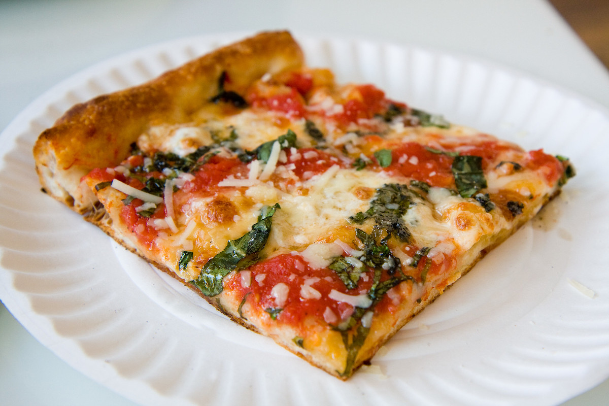 Primer plano de un plato blanco con una porción de pizza de la abuela, inventada en Nueva York. Es una masa delgada en forma cuadrada, cubierta con tomate, queso y albahaca.