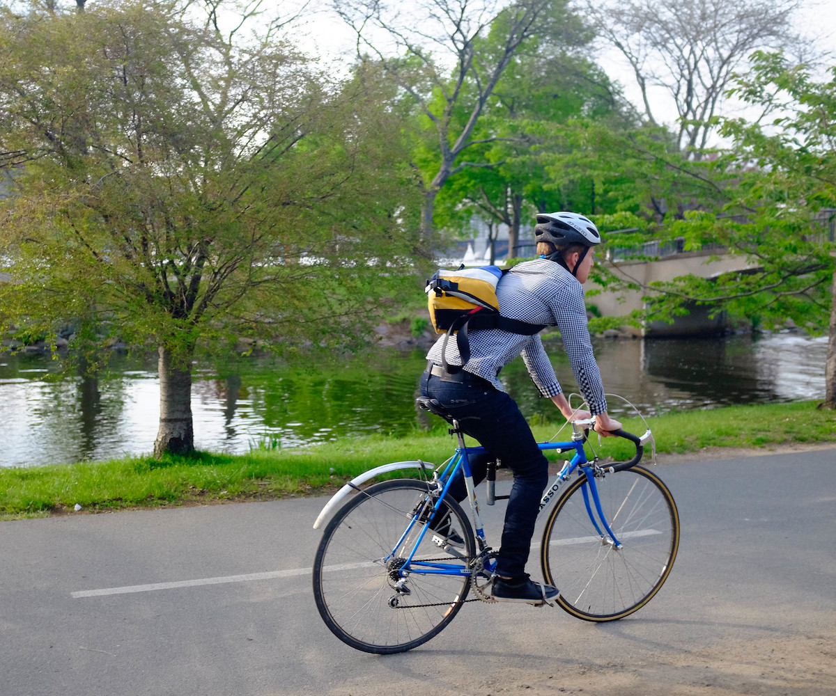 Una persona con jeans, una camisa azul abotonada, una bandolera y un casco monta en bicicleta a lo largo del río Charles en Boston.