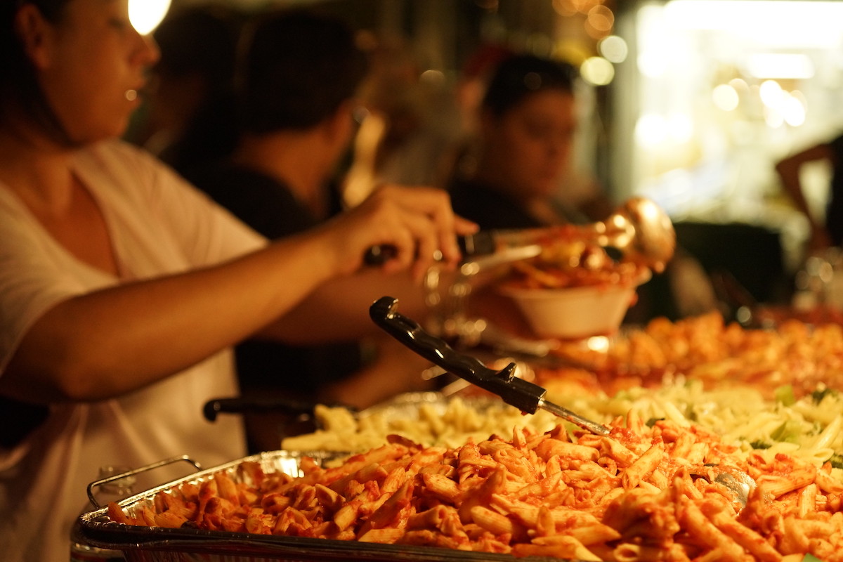Primer plano de una bandeja ziti con gente sirviendo platos de pasta en el fondo. Fisherman's Feast es el festival italiano continuo más antiguo del norte de Boston.