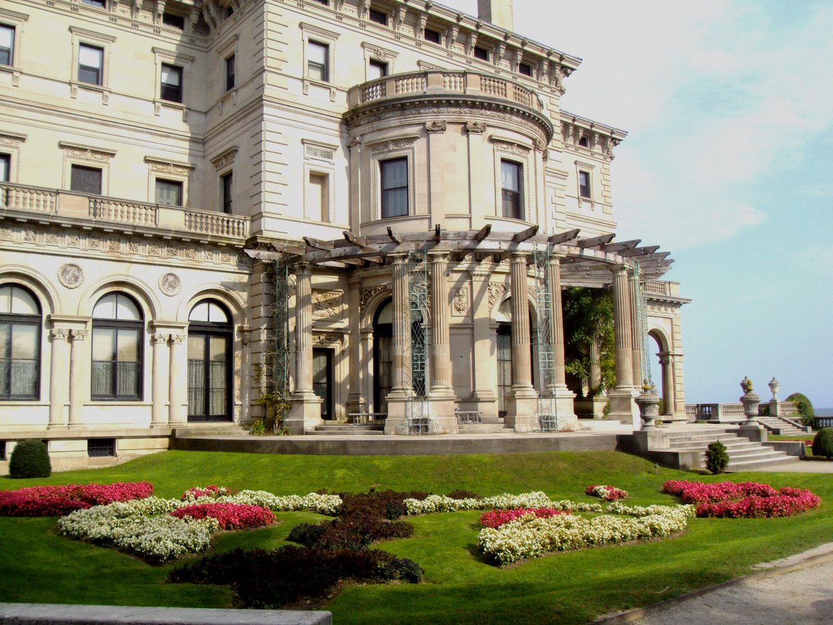 Vista lateral de la mansión Breakers en Newport, Rhode Island. Un gran edificio de piedra blanca con columnas con césped verde sembrado de flores rojas y blancas