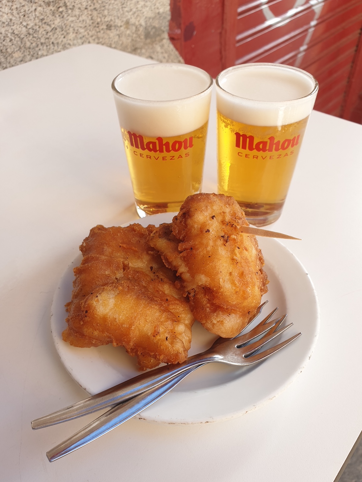 Trozos de bacalao frito en un plato blanco frente a dos pequeños vasos de cerveza.
