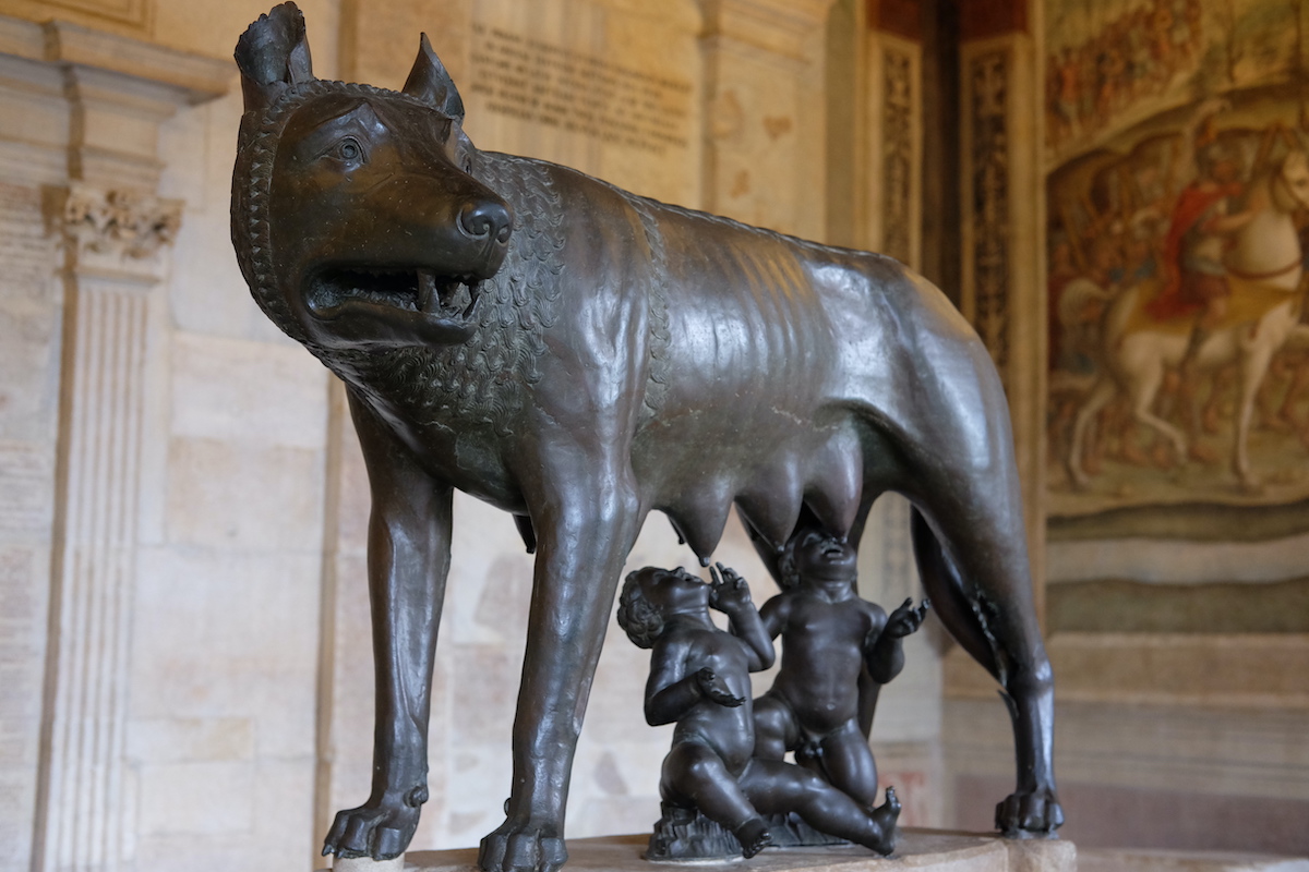 Estatua de bronce de dos pequeñas figuras amamantando debajo de un lobo