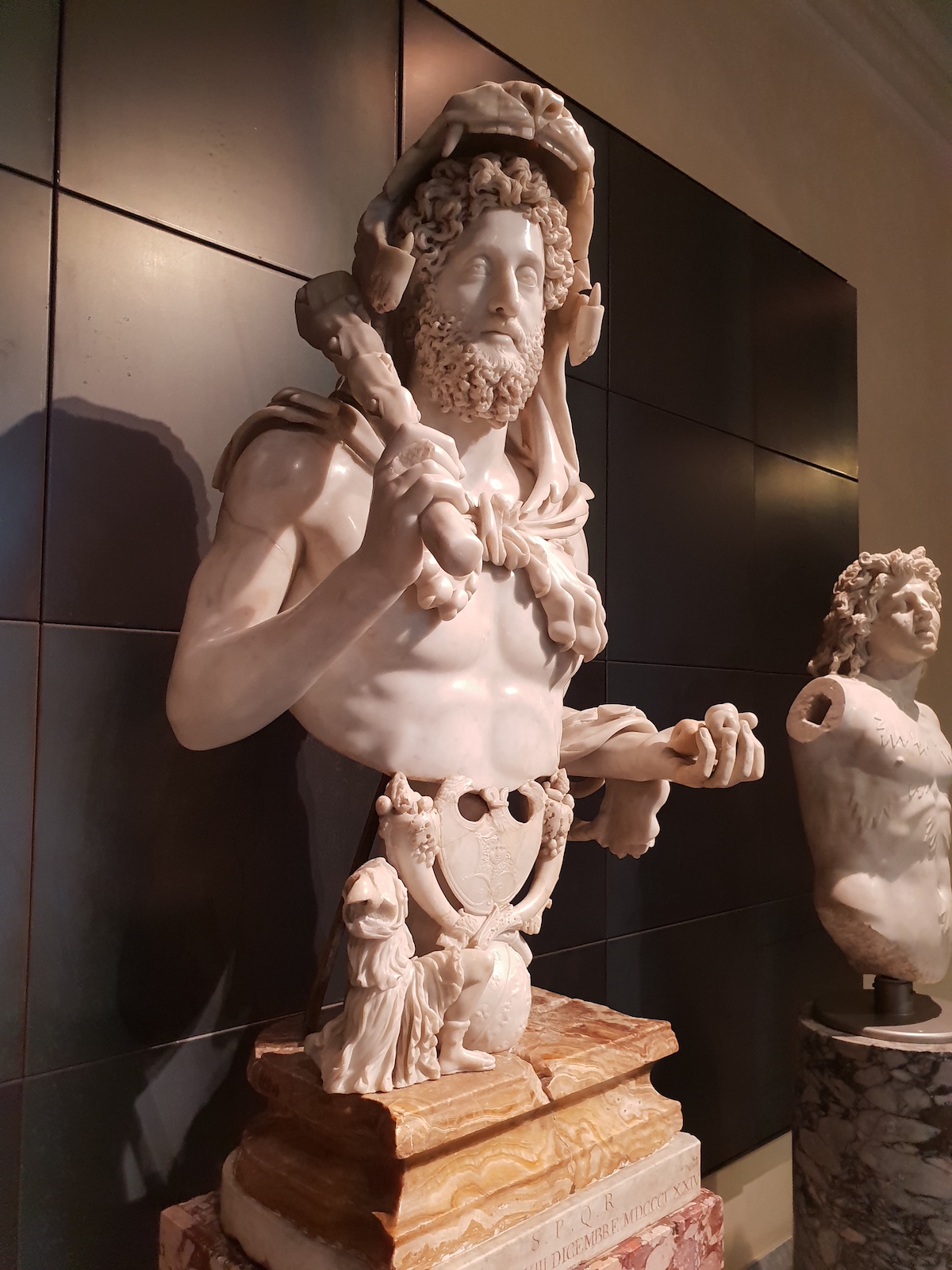 Busto de mármol del emperador romano Cómodo con pelo rizado y barba sosteniendo un garrote sobre su hombro derecho