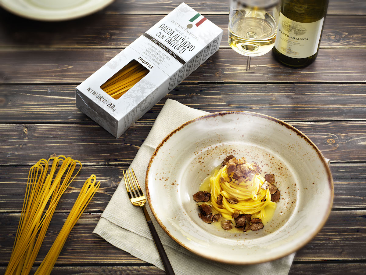 Un plato de pasta con trufa sobre una mesa de madera con una caja blanca de los mismos fideos espaguetis y algunos fideos secos sueltos a la izquierda