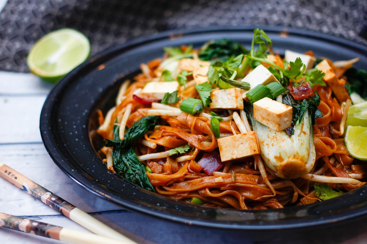 Un plato de padan thai con fideos, tofu, rodajas de lima y cilantro con palillos