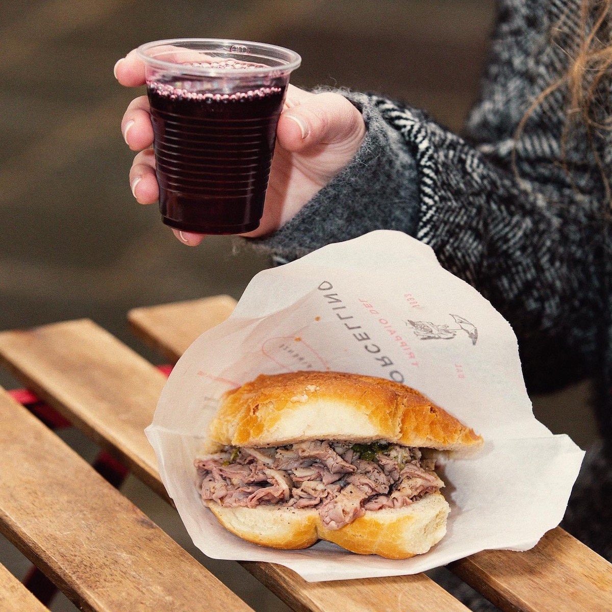 Lampredotto (sándwich de callos) en un envoltorio de papel sobre una mesa de madera con una copa de plástico de vino tinto encima en la mano de una persona