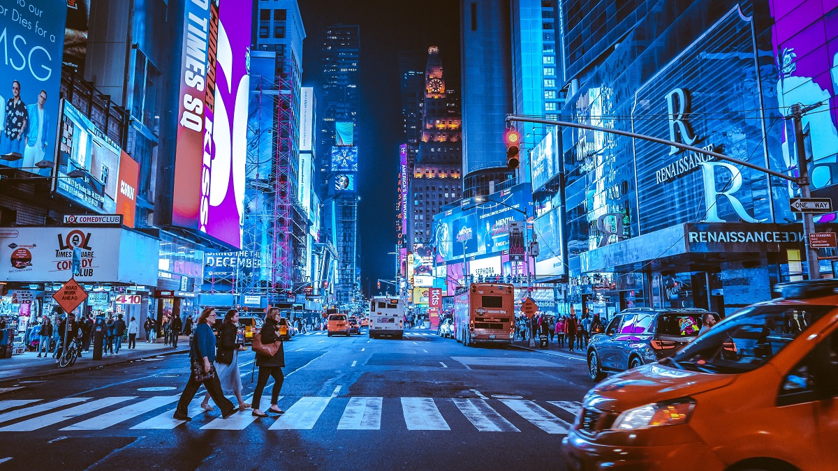 Los peatones cruzan una concurrida intersección de Nueva York por la noche con vallas publicitarias en el fondo