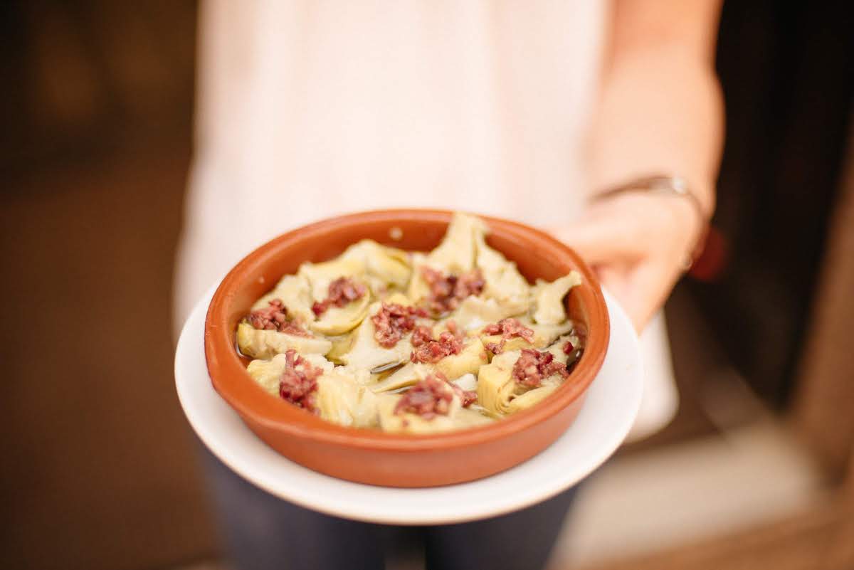 Una mano humana sosteniendo un plato redondo de arcilla de corazones de alcachofa gastados con jamón curado cortado en cubitos.