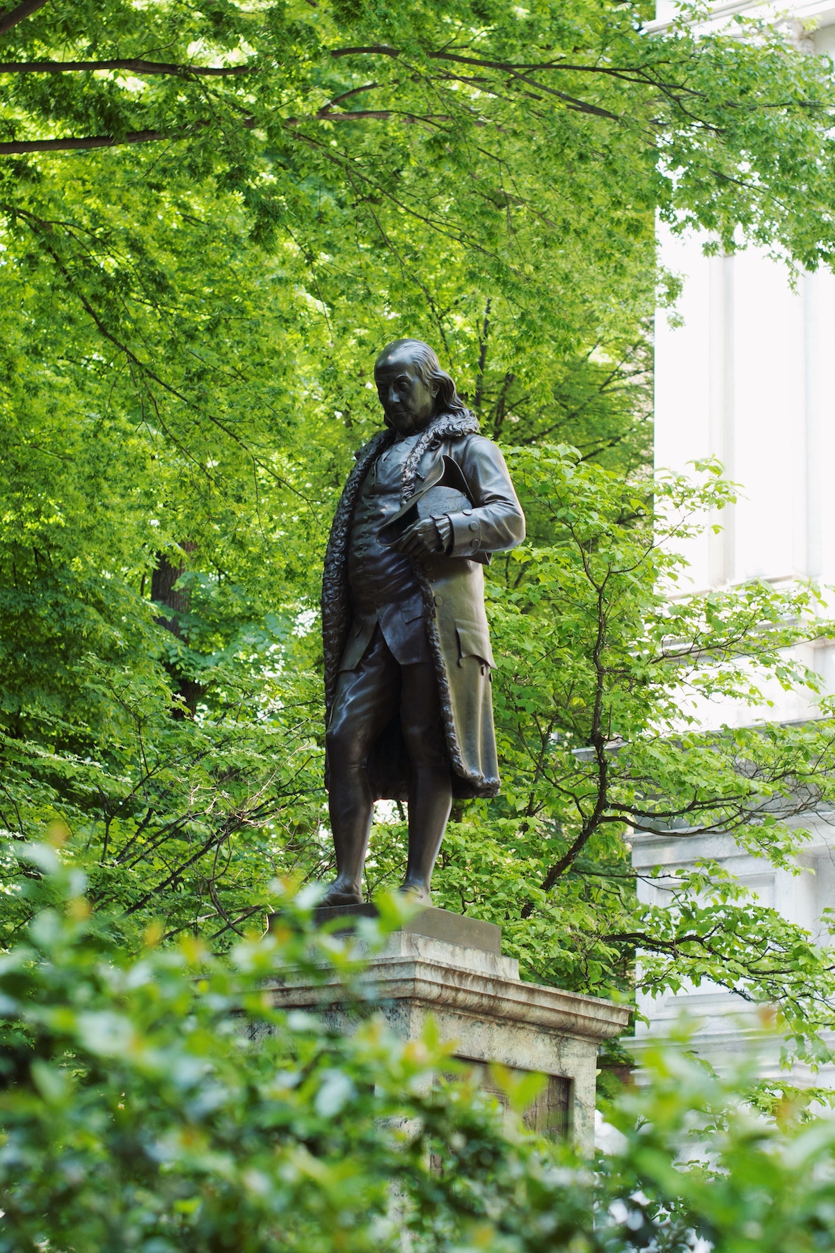 Una estatua de bronce de Benjamin Franklin rodeada de follaje libre brillante