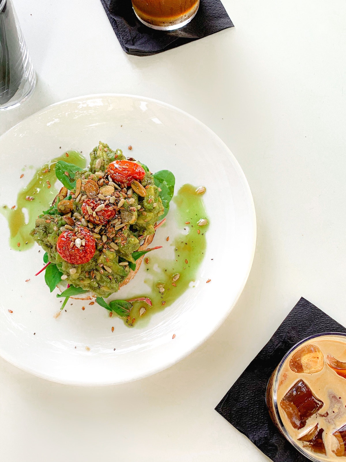 Tostadas de aguacate encima y tomates cherry y pepitas en su plato blanco junto a dos tazas de café helado ligeramente fuera de marco.