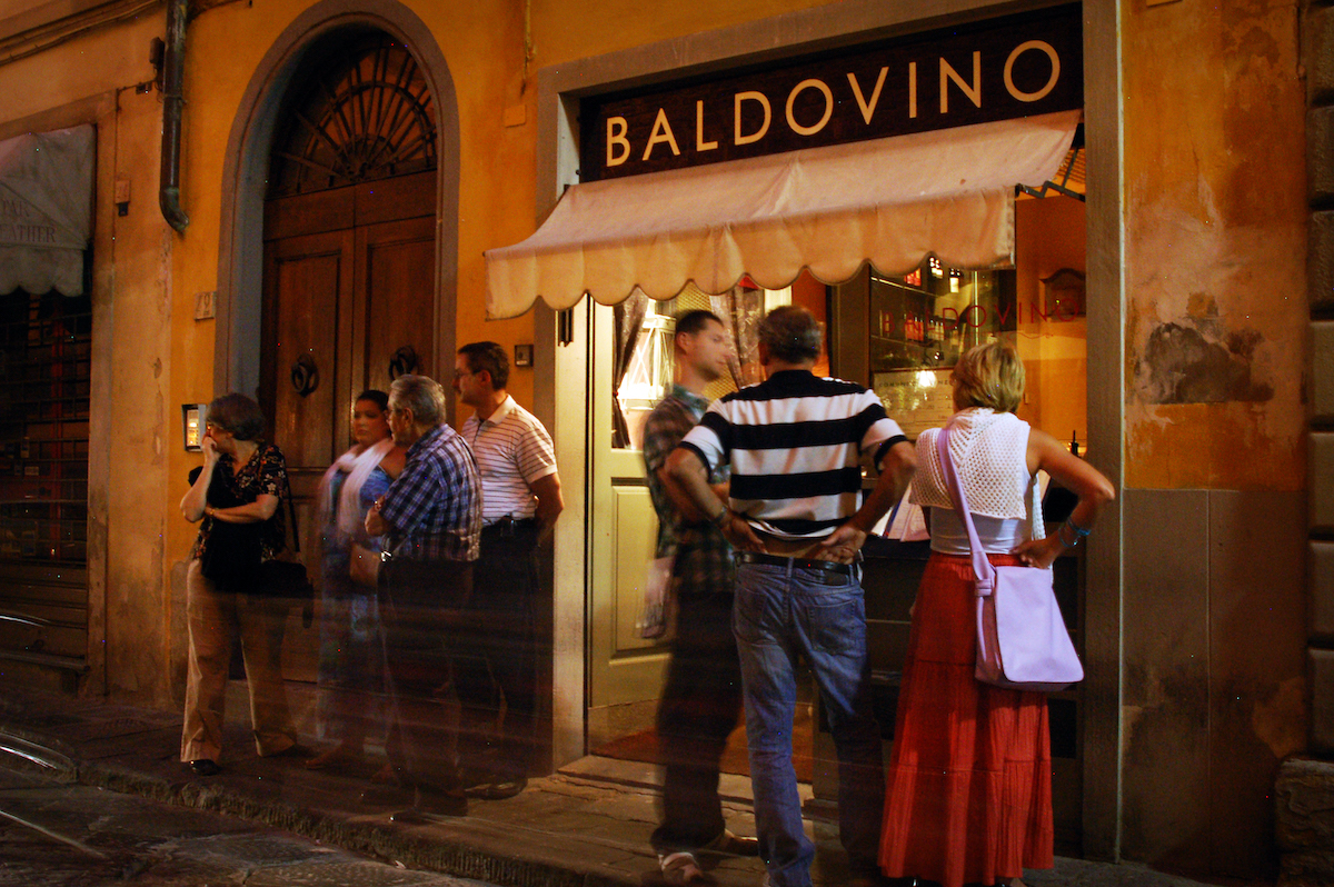 Grupos de personas de pie afuera de un restaurante con el nombre Baldovino sobre el espejo.