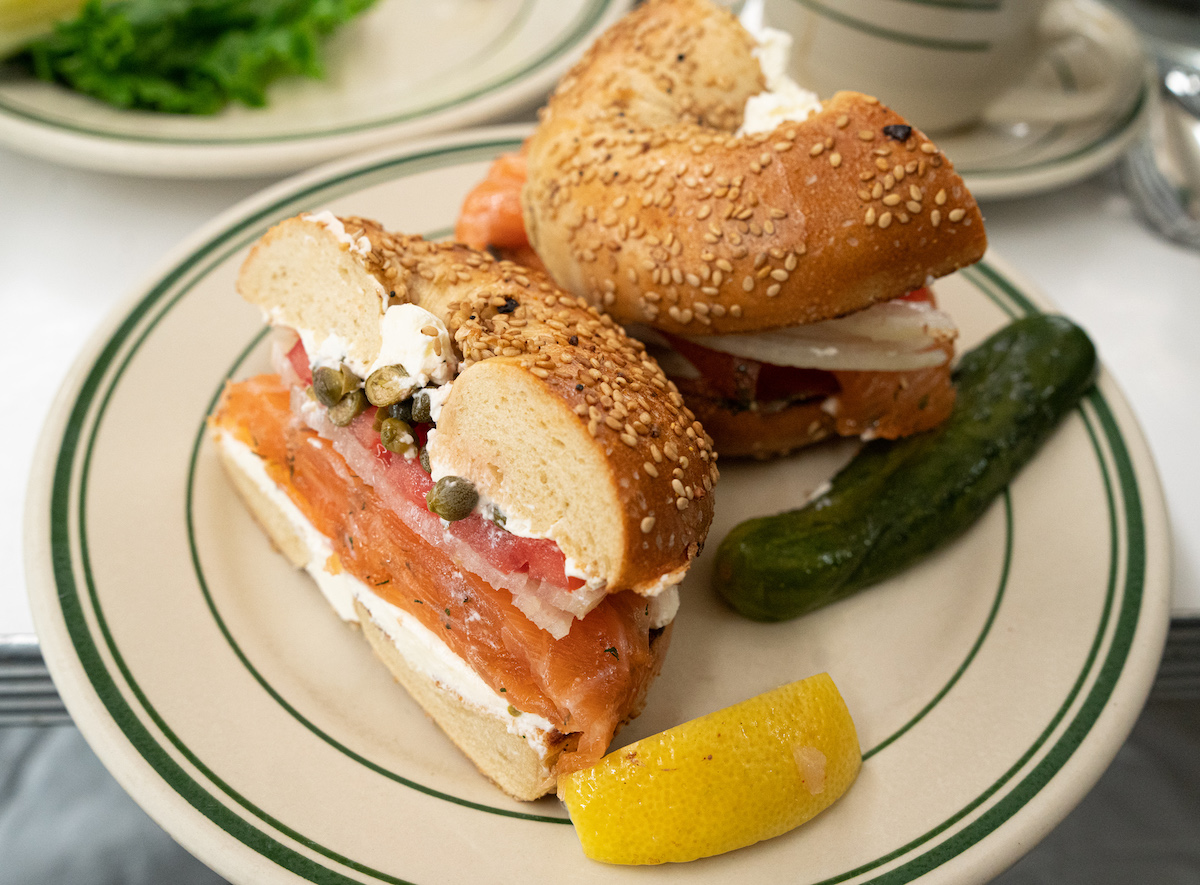 Bagel de queso crema y salmón ahumado en un plato junto a un pepinillo y una rodaja de limón