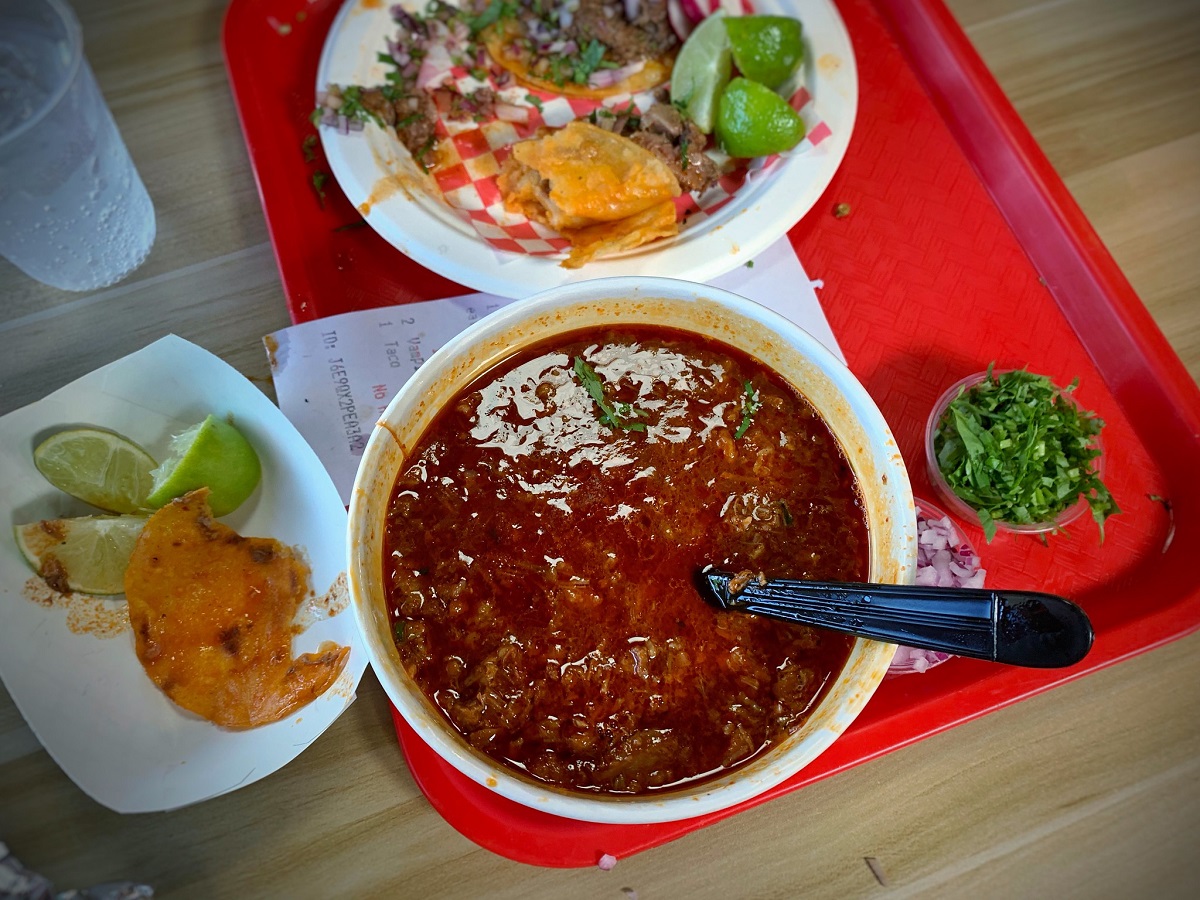 Un plato de abundante sopa roja junto a un plato de tacos en una bandeja roja.