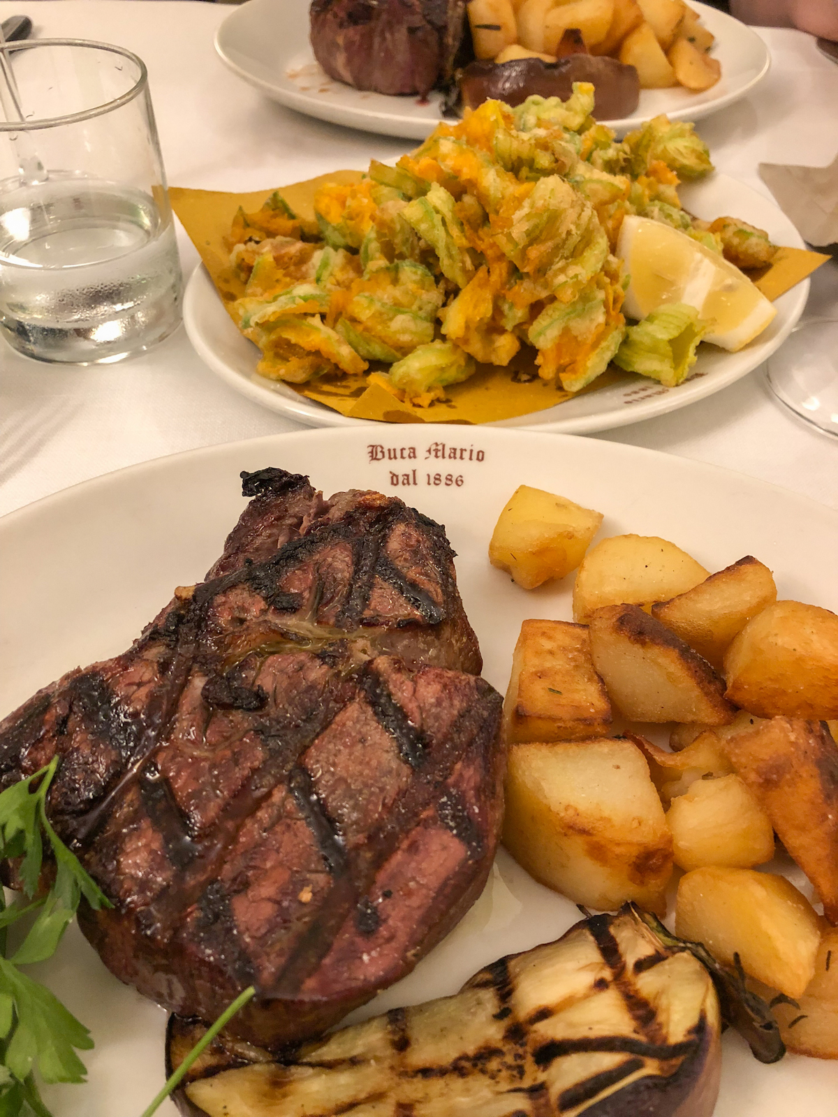 Bistec a la parrilla en un plato junto a papas en cubos, con otros platos visibles en la mesa al fondo
