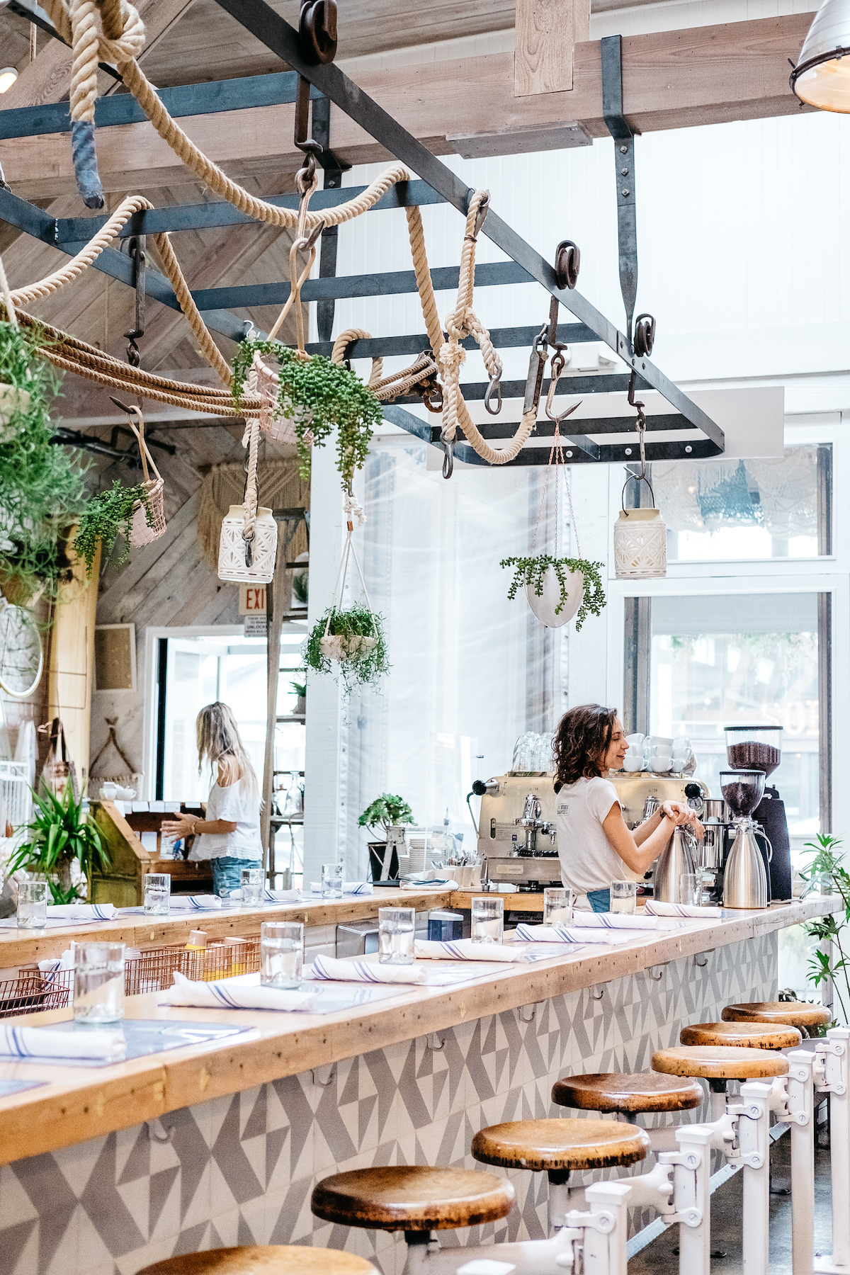 Una zona de bar en un restaurante con grandes ventanales y plantas colgantes encima