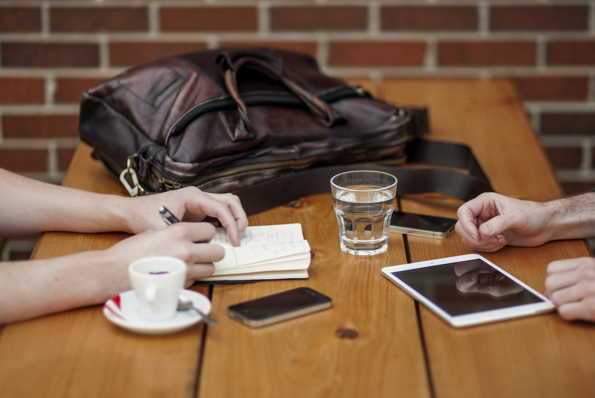 Una mesa de centro de madera con una pequeña taza de espresso en un protector, un pequeño vaso de agua transparente, un teléfono móvil, una tableta, un cuaderno y una mochila