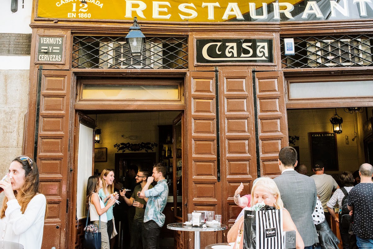 Exterior de un restaurante marrón con gente disfrutando de comidas y bebidas en una terraza al aire libre.