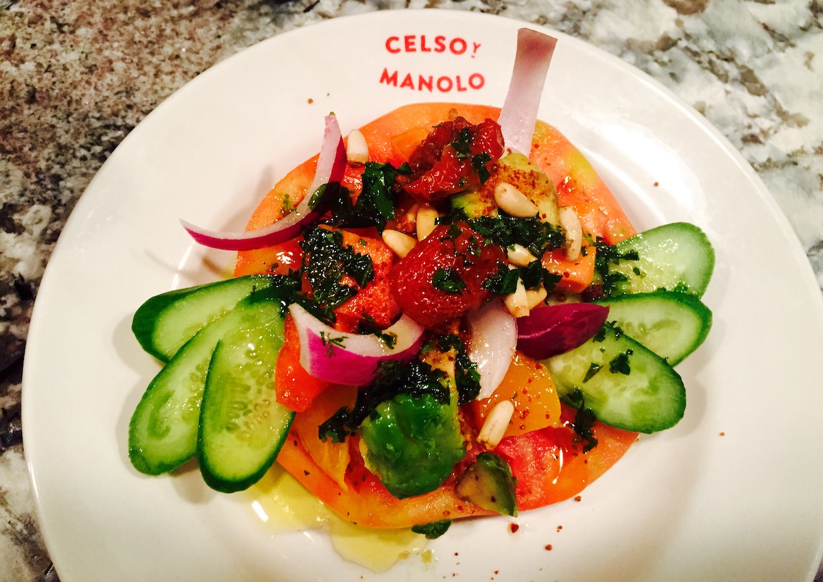 Plato de verduras con base de tomate y guarnición de cebolla roja, pepino, pimiento rojo y hierbas, servido en un plato blanco con la lectura Celso y Manolo