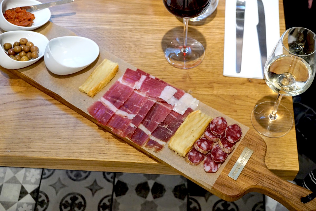 Embutidos y queso expuestos en una tabla de madera junto a dos copas de vino